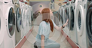 serious girl with long red hair choosing washing machine in appliances store.