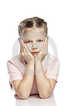 A serious girl with pigtails sits at a table, propping her head with her hands. Isolated over white background.