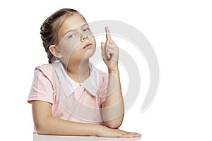 A serious girl with pigtails sits at a table, raising his index finger up. Isolated over white background