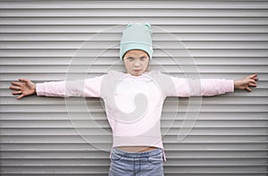 A serious girl in a pink sweater and blue hat stands against a striped wall and spreads her arms wide. Portrait.