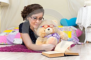 Serious girl and teddy bear reading a book in glasses