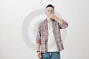 Serious good-looking male model in glasses and trendy checked shirt covering mouth with palm and staring at camera over