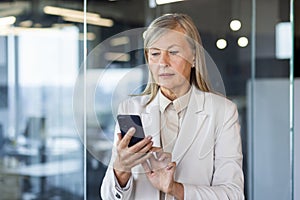 Serious gray-haired businesswoman standing in the office and using the phone, typing and reading messages, making calls