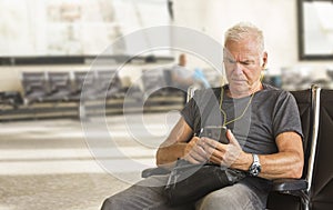 A serious gray-haired man wearing headphones looks at his smartphone while sitting in the departure hall of the airport