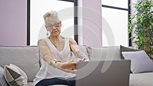 Serious grey-haired senior woman, focused on using laptop, sitting on sofa stretching tired arms at her cozy home living room