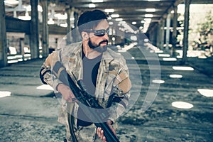 Serious and handsome man in glasses is standing in hangar and holding paintball gun in hands. He is looking straight