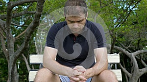 Serious handsome young man sitting in public park with green trees on background