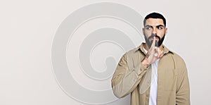 Serious handsome young middle eastern man with beard in casual make shhh gesture with finger lips