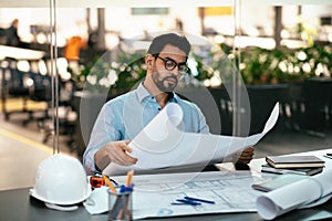 Serious handsome young muslim engineer in glasses with beard with project drawings at workplace with hard hat