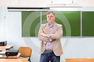 The serious headmaster stands in the classroom at the blackboard with his arms folded