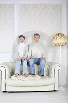 Serious husband and wife sits on back of white leather sofa