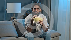 Serious interested Arabic Hispanic Indian bearded man guy wearing glasses 30s male with popcorn watching TV at home sofa