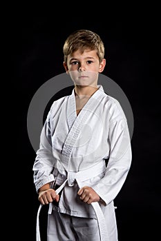 Serious kid in brand new kimono in the front position on the black background. Karate fighter holding his belt