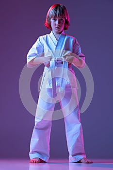 Serious kid, little boy, taekwondo or karate athletes in doboks posing  on very peri color background in neon