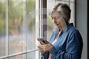 Serious latin female pensioner stand by window texting by cell