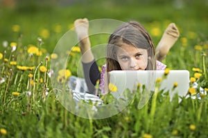 Serious little girl with laptop lying on green grass. Education.