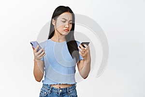 Serious-looking asian girl looking at her credit card, using mobile phone, paying with smartphone, white background