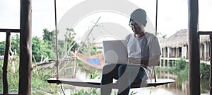 A serious looking Asian man is working hard using his laptop computer. sitting on a wooden swing