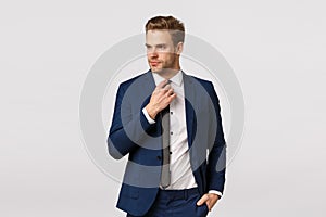 Serious-looking sassy businessman in blue classis suit, adjusting tie and looking away, holding hand in pocket, getting