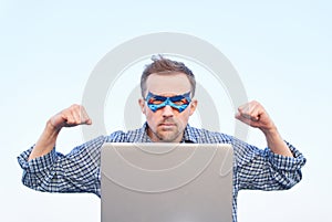 Serious male in blue mask shows strength gesture with laptop and pumped fists