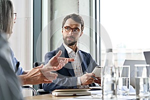 Serious male ceo listen to female colleague holding notebook at table meeting.