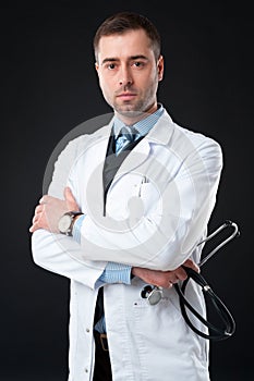 Serious male doctor holds stethoscope in hand isolated on black