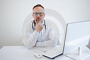 Serious male doctor sits at his workplace in the office and looks at the camera.