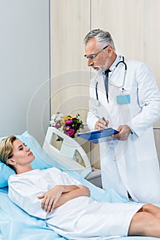 serious male doctor with stethoscope over neck writing in clipboard near adult female patient in hospital