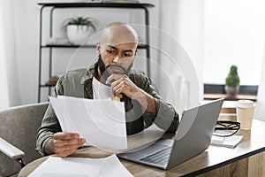 Serious male entrepreneur checking documents, reading financial reports while sitting at desk at home office