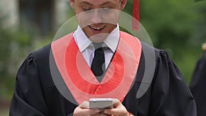 Serious male graduate rubbing chin and looking at smartphone, receive good news