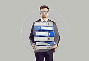 Serious male office worker standing on gray background holding stack of large folders for documents.