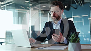 Serious man copying information from smartphone to laptop in office interior