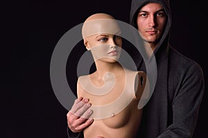 Serious man dressed in black hoodie holding in his arms a plastic women body mannequin on dark background. Loneliness