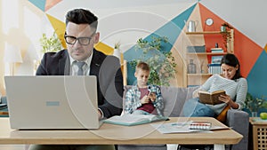 Serious man in formal clothing working from home using laptop, woman and child in background
