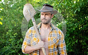 Serious man holding garden spade. Unshaven man wearing cowboy hat and checked shirt. Peasant man