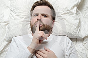 Serious man lies in bed and asks to be silent with his finger to his lips. Silence or secret concept. Top view. Close-up