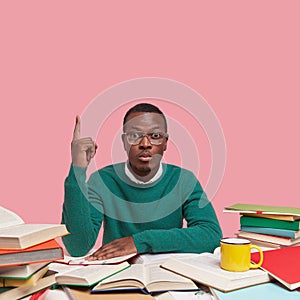 Serious man nerd wears big spectacles, green sweater, points up with one finger, surrounded with many books as prepares