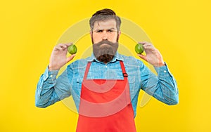 Serious man in red apron holding fresh limes citrus fruits yellow background, fruiterer
