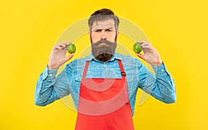 Serious man in red apron holding fresh limes citrus fruits yellow background, fruiterer photo