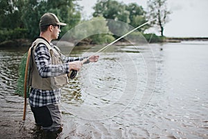 Serious man is standing in water and fishing. He has spinning in hands and fishing net on the back. Guy wears vest