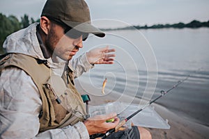 Serious man with sunglasses is sitting at the edge of water and looking down. He is holding spoon with bait. Fly rod is