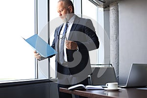 Serious mature business man in formalwear reading contract while standing near the window in the office