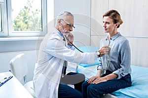 serious mature male doctor examining female patient by stethoscope