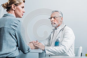 serious mature male doctor holding hands of female patient at table