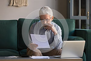 Serious mature man sitting on couch received invoice analyzes expenses