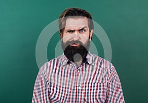 serious mature teacher on background of blackboard. brutal bearded man wear casual checkered shirt and frowns. express