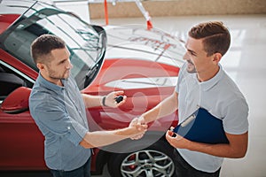 Serious men stand and shake each other`s hands. Purchaser holds car key. Dealler hold tablet. They made a deal. Red car