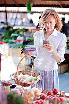 Serious mid adult woman buying vegetables in farmer`s market, looking at shopping list on her mobile phone