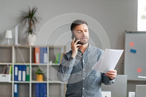 Serious middle aged businessman working at office, standing by workdesk, holding documents and having phone conversation