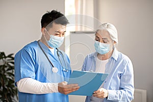 Serious middle aged korean doctor in protective mask giving contract sign to old caucasian female patient